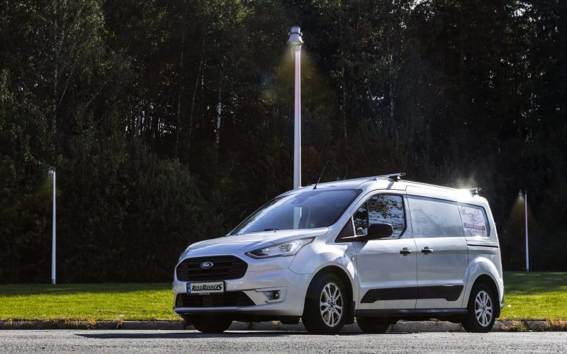 Liten hvit budbil med en Road Runners logo på siden, med masse trær og lys i bakgrunnen.