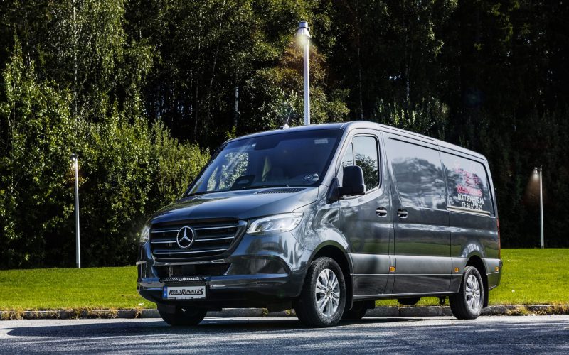 Fronten av en mørkegrå budbil med Road Runners logoen på siden. Masse trær og lys i bakgrunnen.