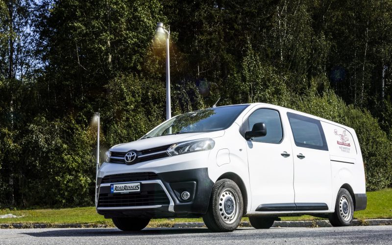 Hvit budbil med en Road Runners logoen på siden, med masse trær og lys i bakgrunnen.