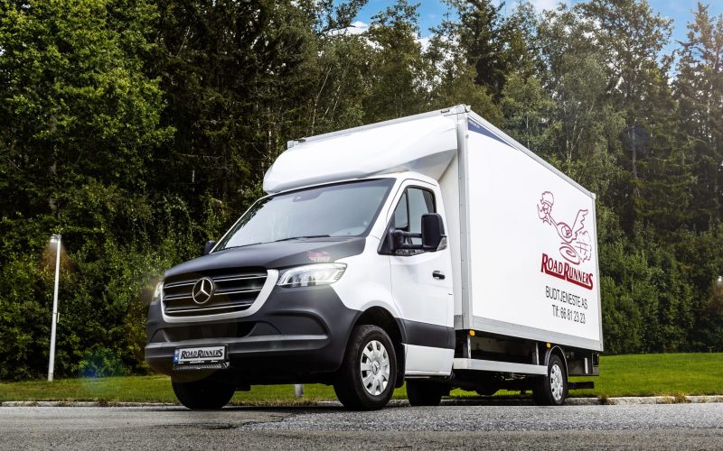 Hvit budbil med en Road Runners logoen på siden, med masse trær i bakgrunnen.